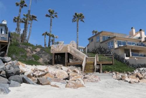 A sandy beach with large rocks, palm trees, and a wooden staircase leading up to a modern house.