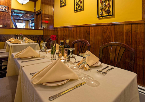 Cozy restaurant interior with neatly set tables, beige tablecloths, and decorative wall art. A rose centerpiece adds charm.