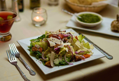 A fresh salad with lettuce, tomatoes, onions, and cheese on a white plate, accompanied by a candle and side dishes.