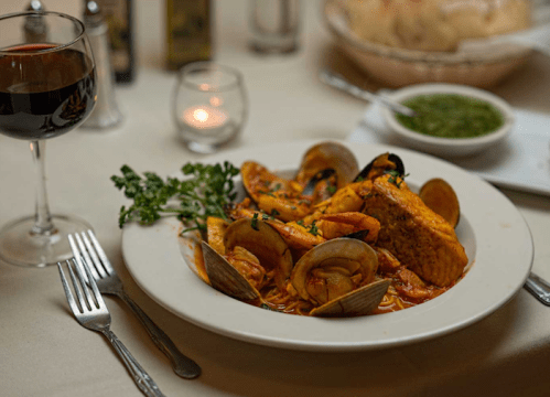 A plate of seafood pasta with shrimp, clams, and salmon, accompanied by a glass of red wine and a candlelit setting.