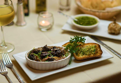 A dish of mushrooms in sauce, garnished with herbs, served with toasted bread and a glass of white wine.