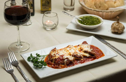 A plate of cheesy baked pasta in marinara sauce, accompanied by a glass of red wine and a side of green sauce.
