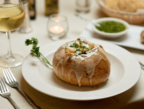 A bowl of creamy soup served in a bread bowl, garnished with herbs, alongside a glass of white wine.