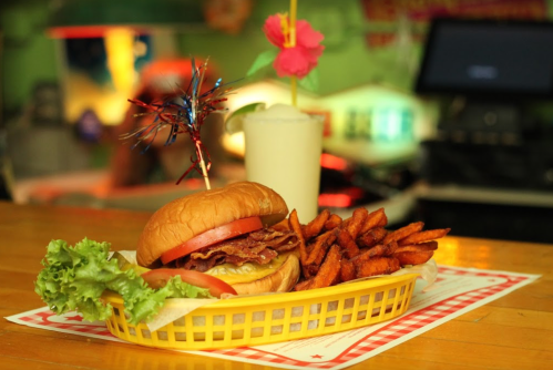 A delicious burger with bacon, lettuce, and tomato, served with sweet potato fries and a tropical drink in a basket.