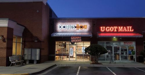 A storefront at dusk featuring "DONUTS NOW" with a "GRAND OPENING" sign and "U GOT MAIL" next door.