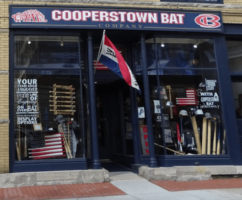 Storefront of Cooperstown Bat Company featuring wooden bats, flags, and a sign indicating it's open.