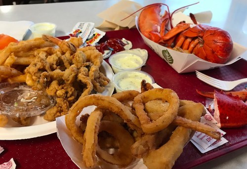 A tray of fried seafood, including onion rings, calamari, and a lobster, accompanied by dipping sauces.