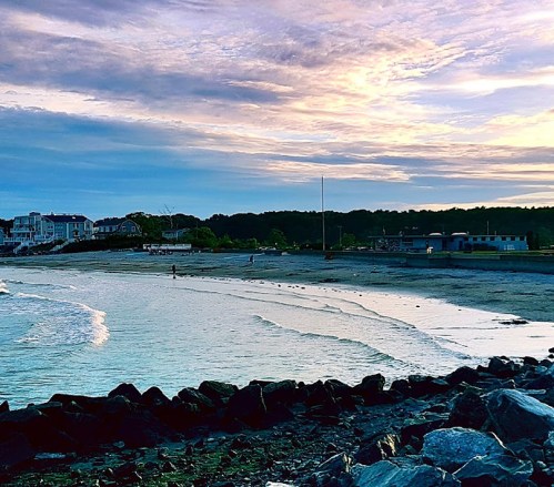 A serene beach scene at sunset, with gentle waves, rocky shore, and distant figures walking along the water's edge.