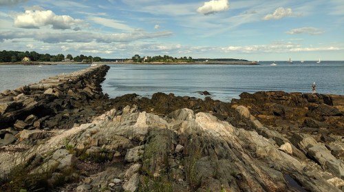 A rocky shoreline leads to a calm sea with distant sailboats and a scenic sky, creating a peaceful coastal view.