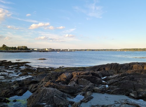 A serene coastal scene featuring rocky shores, calm waters, and a clear blue sky with scattered clouds.