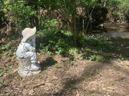 A white statue of a child in a straw hat sits on a stump, gazing at a serene stream surrounded by greenery.