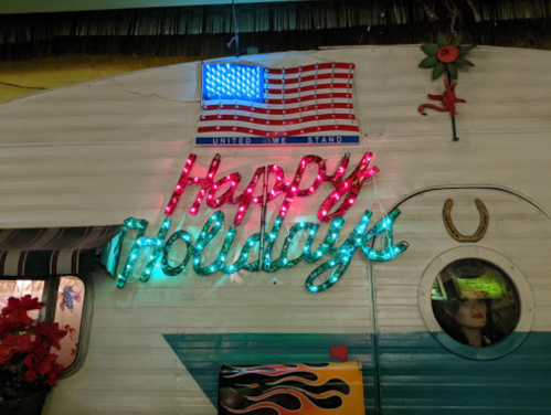 A colorful sign reading "Happy Holidays" with an American flag above it, displayed on a vintage trailer.