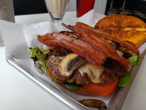 A close-up of a burger topped with bacon, mushrooms, and cheese, served with a side of a toasted bun and a milkshake.