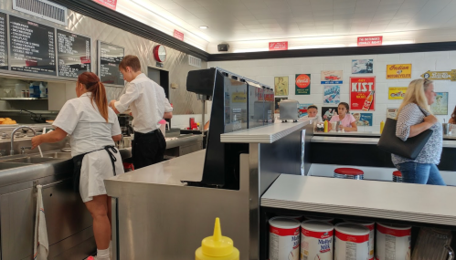A retro diner scene with staff at the counter, customers seated, and vintage advertisements on the walls.