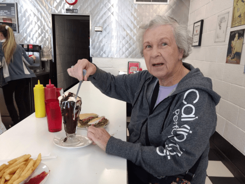 An older woman enjoys a sundae at a diner, with fries and a sandwich on the table in front of her.