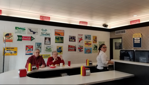 A diner scene with two patrons at a counter, a waitress serving, and vintage advertisements on the walls.