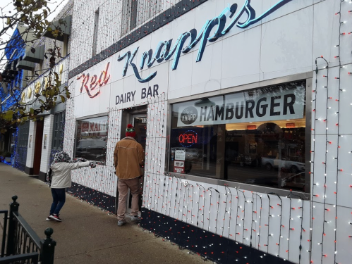 A child and an adult approach the entrance of Red Knapp's Dairy Bar, decorated with holiday lights.