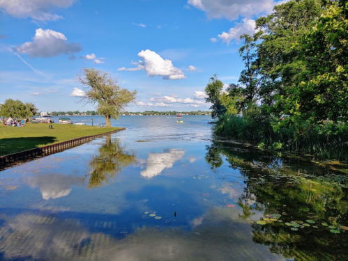 A serene lake scene with blue skies, fluffy clouds, and reflections on the water, surrounded by trees and greenery.