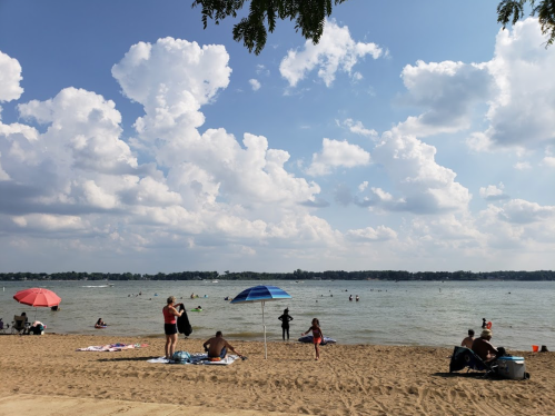 A sandy beach with people sunbathing, swimming, and colorful umbrellas under a partly cloudy sky.