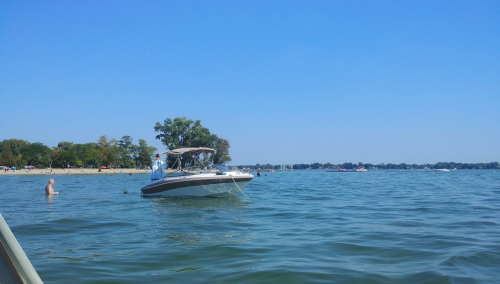 A boat on a calm lake with a person swimming nearby and a beach in the background under a clear blue sky.