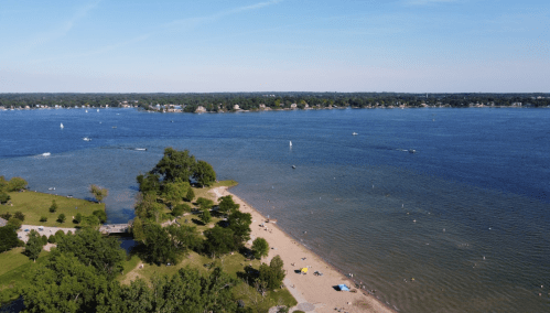 Aerial view of a beach with people swimming, boats on the water, and green trees along the shoreline.