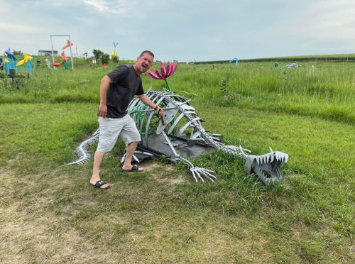 A man in a black shirt and sandals poses next to a large, artistic skeleton of a fish in a grassy field.