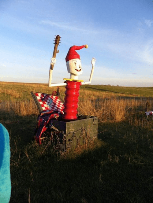 A whimsical sculpture of a smiling figure with a jester hat, holding a stick, set in a grassy field.