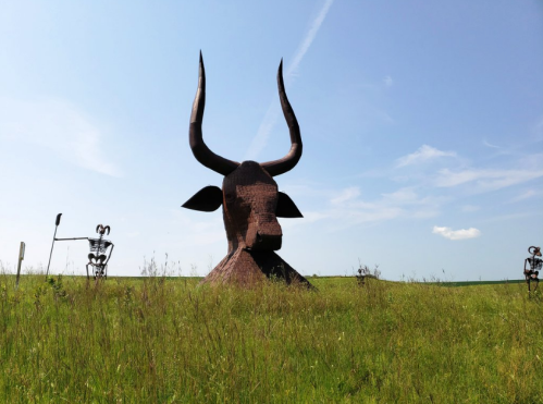 A large metal bull sculpture stands in a grassy field, with two smaller skeletal figures nearby under a blue sky.