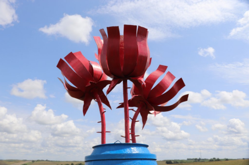 Three large red metal flowers atop a blue base, set against a backdrop of a cloudy sky.