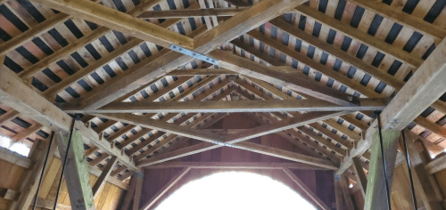 Interior view of a wooden barn, showcasing exposed beams and a sloped roof with a bright light at the entrance.