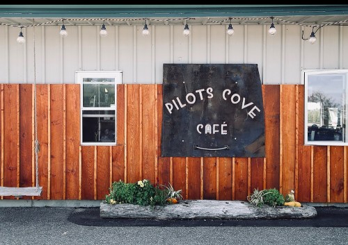 A rustic café exterior with a wooden wall, a sign reading "Pilots Cove Café," and string lights above.