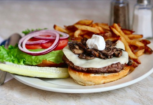 A juicy burger topped with mushrooms and cheese, served with fries, lettuce, tomato, onion, and a pickle on the side.
