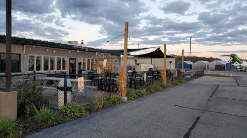 Outdoor dining area with tables and chairs, surrounded by a fence, under a cloudy sky near a building and hangars.