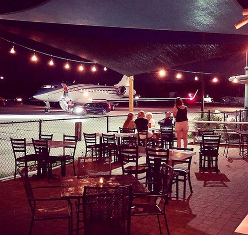 A nighttime scene at an outdoor restaurant with tables, overlooking a private jet on the runway.