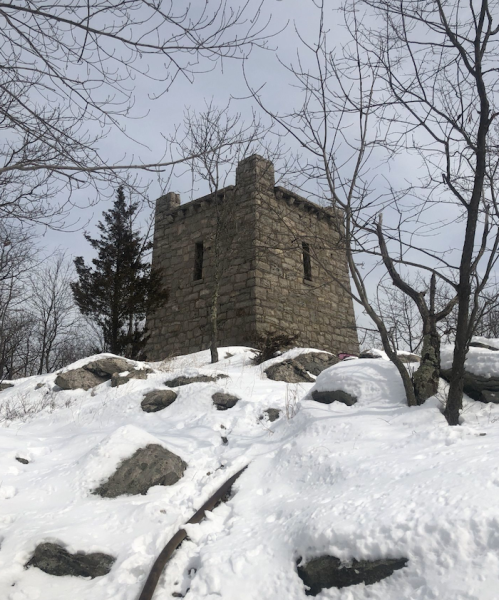 A stone tower stands on a snowy hillside, surrounded by bare trees and large rocks under a cloudy sky.