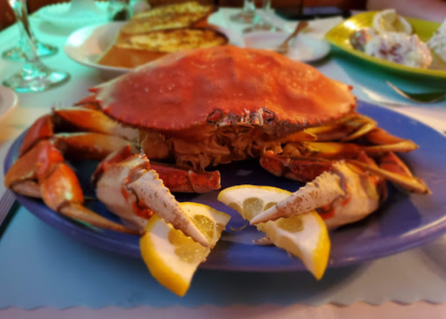 A large cooked crab on a blue plate, garnished with lemon wedges, surrounded by seafood dishes in the background.