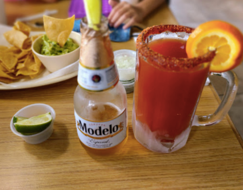 A refreshing drink with an orange slice and a beer bottle, accompanied by chips and guacamole on a wooden table.