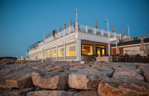 A seaside restaurant with a deck, surrounded by large rocks, under a clear blue sky at dusk.