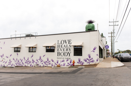 Exterior of Thistle Farms with a mural reading "LOVE HEALS EVERY BODY" and purple floral designs on the wall.