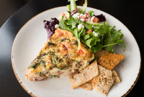 A slice of frittata with greens and tomatoes, served with a side salad and crispy crackers on a white plate.