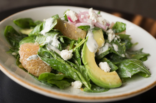 A fresh salad with spinach, avocado, grapefruit, red onion, and a creamy dressing, served in a white bowl.