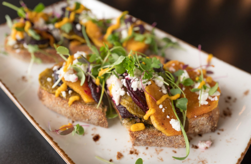 A plate of colorful open-faced sandwiches topped with roasted vegetables, microgreens, and drizzled with sauce.