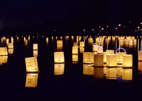 Floating lanterns illuminate a dark lake, reflecting warm light against the water's surface.