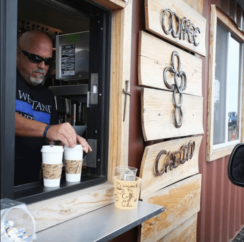 A barista hands two coffee cups to a customer at a coffee stand with a wooden sign.