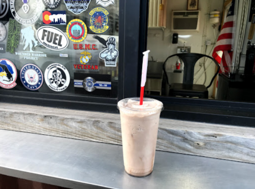 A milkshake with a red straw sits on a counter in front of a window covered in various stickers and an American flag.