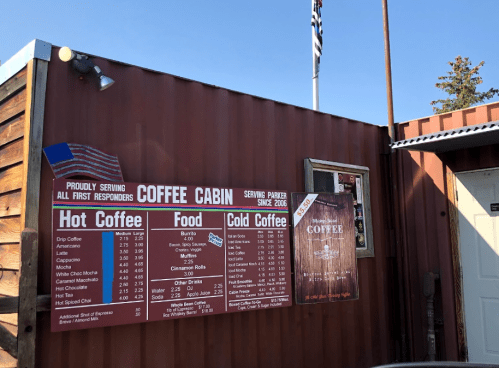 A wooden coffee stand menu displaying hot and cold coffee options, food items, and prices. American flag in the background.