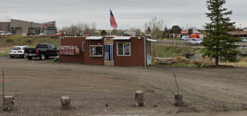 A small, rustic building with an American flag, surrounded by gravel and trees, near a road and parking area.