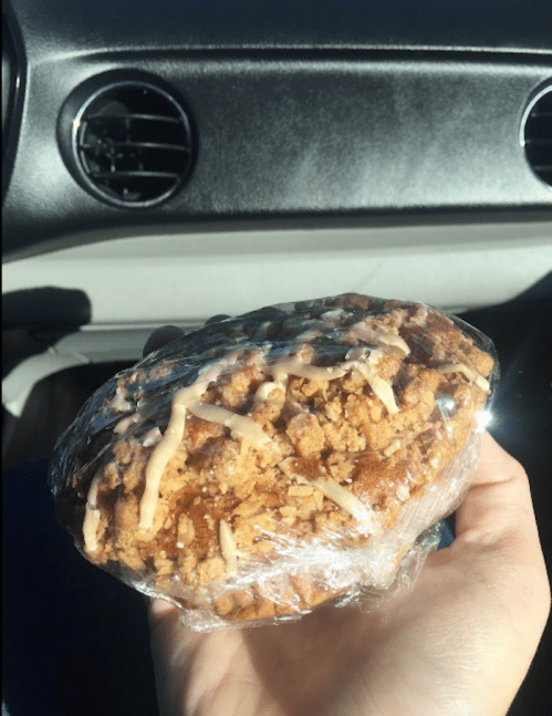 A hand holding a large, wrapped pastry topped with crumbly topping and drizzled icing, inside a car.