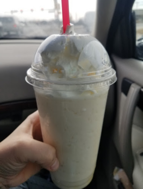 A hand holding a large milkshake topped with whipped cream and a red straw, inside a car.