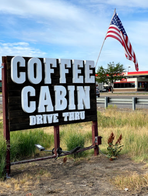 Sign for "Coffee Cabin" with a drive-thru, featuring an American flag in the background and grassy surroundings.